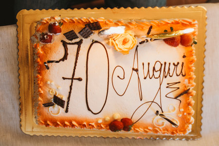 Decorative cake celebrating a 70th birthday features chocolate pieces, strawberries, and a floral accent. Script reads Auguri, adding an Italian touch to this special occasion dessert.の写真素材