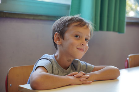 Young boy sitting at a school desk with hands folded, wearing a content expression. The background...