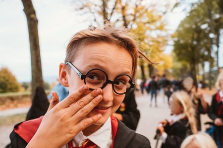 Child dressed as a wizard playfully adjusts glasses with a smile during a park gathering, surrounded by others in similar attire. The scene captures a lighthearted momentの写真素材