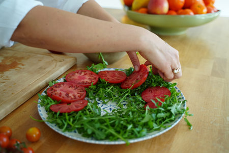 Fresh tomato slices and arugula are arranged on a decorative plate, showcasing a vibrant salad preparation. A hand slices a tomato on a wooden board, adding freshness to the scene.の写真素材