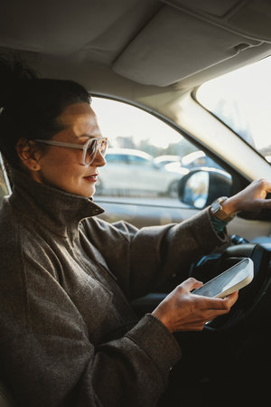 Woman in car holding smartphone while driving through traffic. She is focused and concentrated, indicating multitasking and use of technology while commuting in a busy urban area.の写真素材