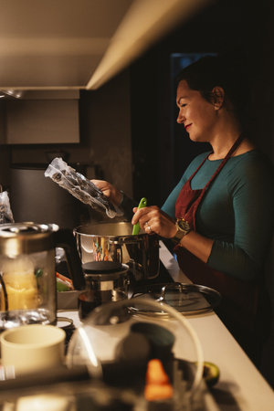 Woman smiling while using sous vide technique in the kitchen. She prepares food with focus and enjoyment, showing modern culinary methods. The scene captures a blend of tradition and innovation.の写真素材