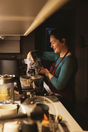 Woman adds fresh mussels to a pot on the stove, preparing a seafood dish. The kitchen setting features various cooking utensils, emphasizing culinary activity and home cooking.の写真素材