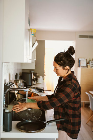 Woman wearing a plaid shirt washes a glass lid in a modern kitchen, focusing on household chores. The kitchen setup includes various utensils and a clean, tidy environment.の写真素材