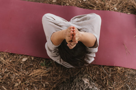 Individual practicing a seated yoga pose outdoors on a mat, with hands in prayer position overhead....