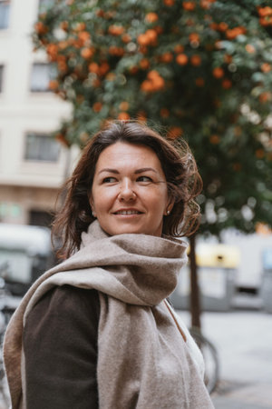 Woman in cozy scarf smiling outdoors with orange trees and buildings in the background. Hair gently...