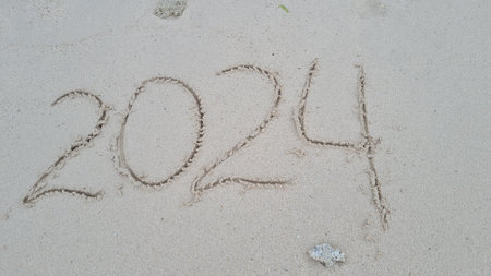 2024 hand written in sand on a beautiful beach. Textured sand with footprints and water.