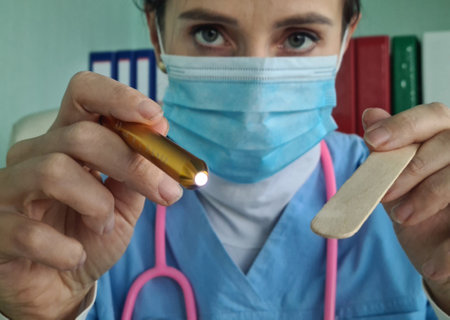 Female otolaryngologist shines light and holds spatula into throat of patient with pain syndromes....