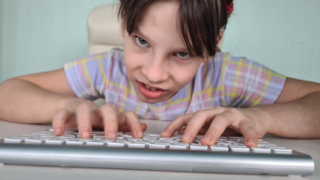 A young girl with pigtails is sitting at a wooden desk and typing on a laptop keyboard. She has an amused expression on her face, as if she is sticking her tongue out playfully.の写真素材