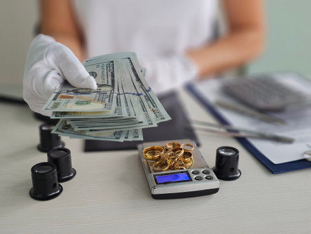 A person wearing white gloves hands over cash while a scale and gold jewelry sit on a desk at a jewelry appraisal facilityの写真素材