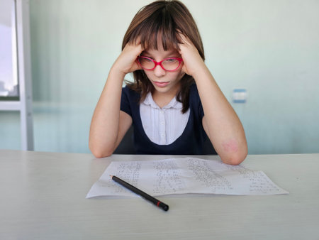 Schoolgirl with glasses looking upset and tired while working on homework at desk in classroom. Difficult homework and stress and tiredness from studyingの写真素材