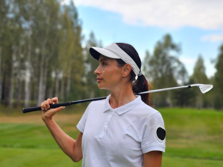 Golf player poses on the course while holding a club against a backdrop of trees and open grass under a partly cloudy skyの写真素材