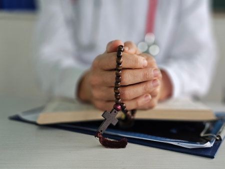 Praying hands clasped over a book with a rosary in a serene setting during a moment of reflectionの写真素材