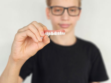 Young man holding a set of artificial teeth and showing dentures. Bite alignment teeth whitening in...