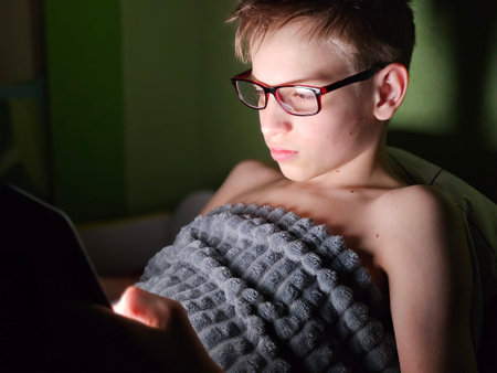 A boy reading on a tablet while covered with a cozy blanket in a dimly lit room in the eveningの写真素材