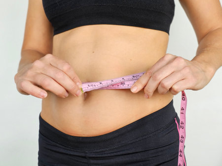 A woman measures waist with a tape measure while wearing athletic clothing in a well-lit indoor...