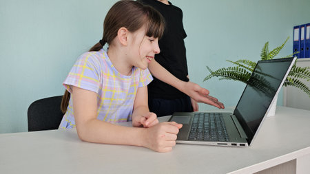 Young girl engaged in learning at a desk while another child assists her with a laptop in a bright...