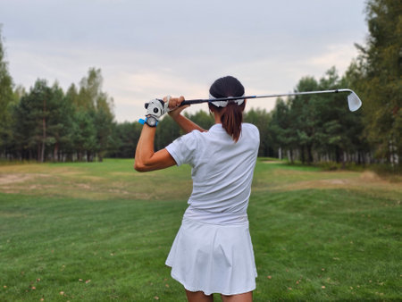 A female golfer swings her club on a lush green fairway surrounded by trees during a cloudy day at the golf courseの写真素材