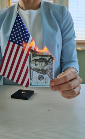 Person holds a burning hundred-dollar bill near a small American flag in a well-lit indoor settingの写真素材