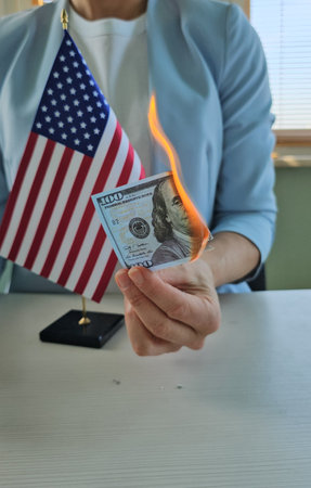 A person is holding a dollar bill that is on fire while standing beside a small American flag