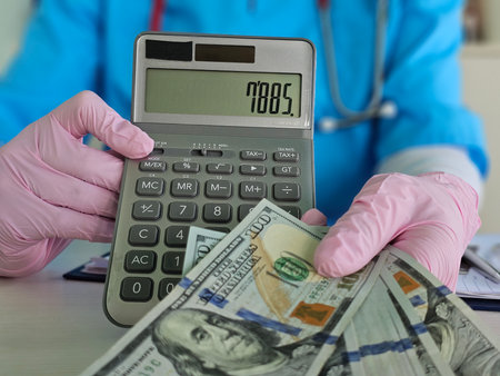 Healthcare professional calculates expenses while handling cash and using a calculator in a clinical settingの写真素材