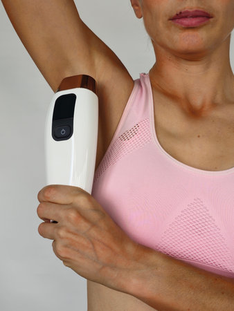 Woman using a hair removal device on her underarm while wearing a pink sports bra during a personal...