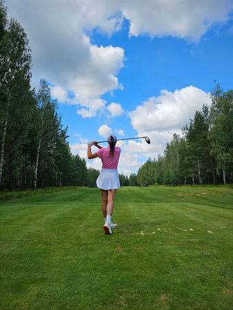 Golfer swinging club on a green course under a blue sky with clouds in late afternoonの写真素材