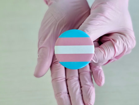 Hands in gloves hold a circular pin featuring the transgender pride flag colorsの写真素材