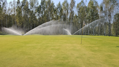 Watering the green during a sunny day at a golf course with trees in the backgroundの写真素材