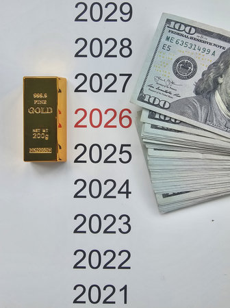 A gold bar is placed next to a stack of cash, with a calendar showing the years 2021 to 2029, focusing on 2026.の写真素材