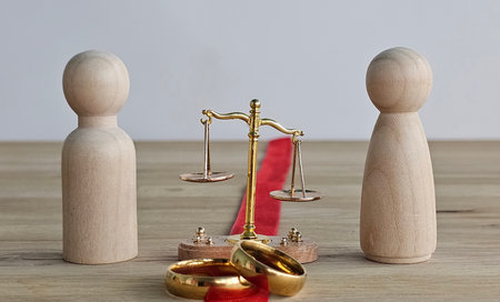 Figures stand beside scales of justice and wedding rings on a wooden surface with a red ribbon in a neutral settingの写真素材