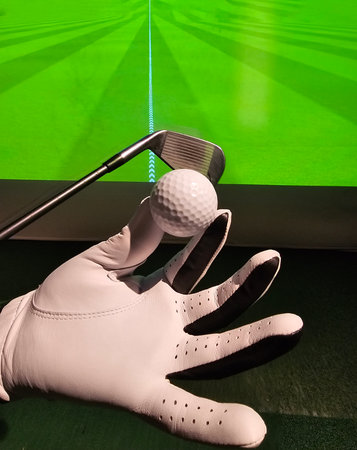 Golf club held by a gloved hand with a golf ball in front of a green backdrop showing a courseの写真素材