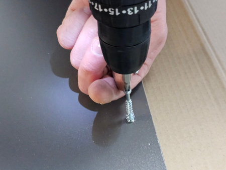 Hand uses a drill to fasten a screw into a flat surface in a workshop during daylight hours