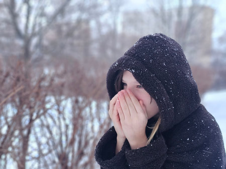 Child covers face in snow while standing outside in winter and surrounded by trees and bushes during...