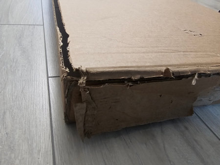 Cardboard box rests on a wooden floor showing wear and damage from handling and movement over time