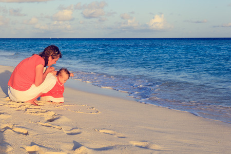 mother and little daughter drawing heart on sand beachの写真素材