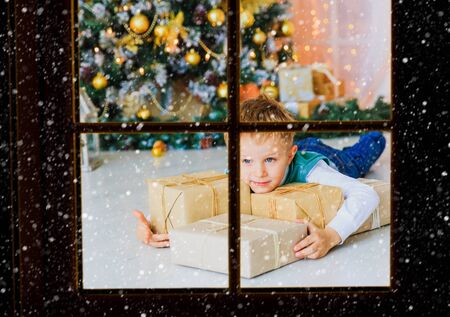 little boy with christmas presents in decorated living room, view from outside windowの写真素材
