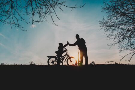 silhouette of happy father and son riding bike and scooter in sunset natureの写真素材