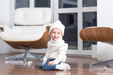 Child sits at the window on the floor. Portrait of a girl in a white hat and sweater.の写真素材