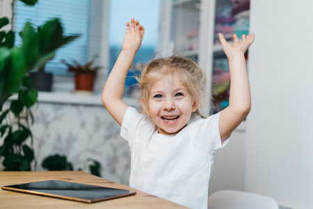 a little girl is sitting at a table with a tablet with her hands raised in the air smiling and...
