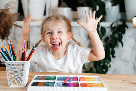 a little blonde happy girl laughs, shows her tongue and sits at a table with a bright colorful...