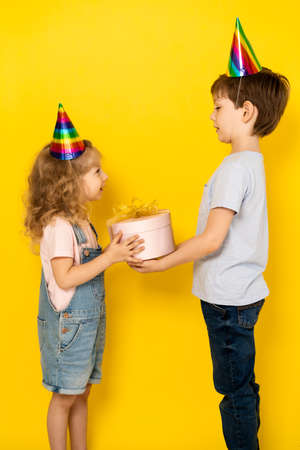 Little blonde girl accepts gift in pink box for her birthday from her brother, on a yellow...