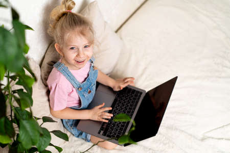 Happy little blonde girl sits at home on a bed with a laptop, a view from above, a place for text,...