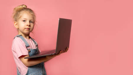 A smiling little girl stands with a PC laptop in her hands, isolated on a pink background. School distance education at home during quarantine concept. banner with space for textの写真素材