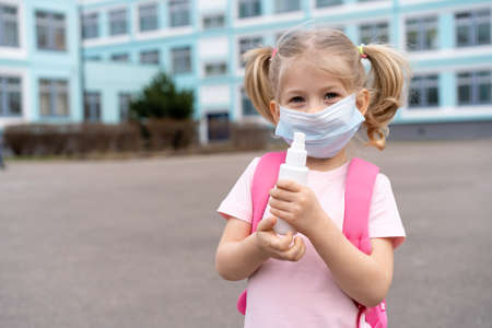 a little girl near the school with a backpack in a mask and an antiseptic in her hands, the concept of virus preventionの写真素材