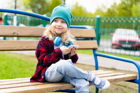 Little blonde girl with smartphone hands and headphones, sitting on a bench in the parkの写真素材