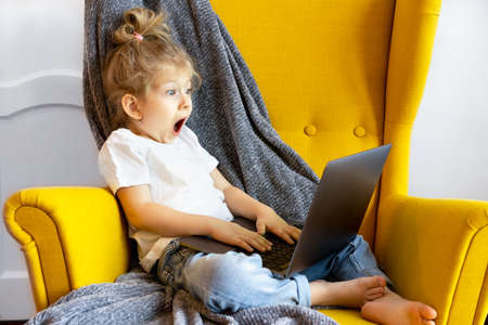a small blonde girl looks at her laptop with a very surprised and frightened look sitting in a chair at homeの写真素材