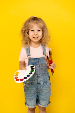 A blonde curly-haired little girl on a yellow background smiles, with paints and brushes in her hands, dressed in a denim jumpsuit and a pink T-shirt, vertical photoの写真素材