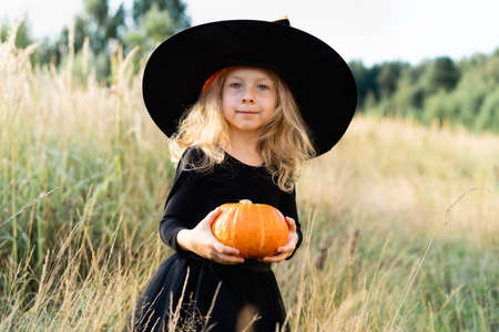 little blonde girl in a black costume and a witch hat, Halloween with a pumpkin in her hands, a cheerful childの写真素材