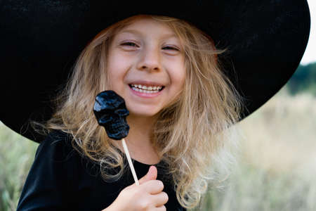 a little blonde girl in a black costume and a witch hat, Halloween with a candy skull in her hands, a cheerful child, close-upの写真素材